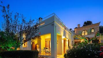 Um edifício moderno de dois andares com varanda, área de jantar com cadeiras e mesa, e um jardim com flores e uma árvore.