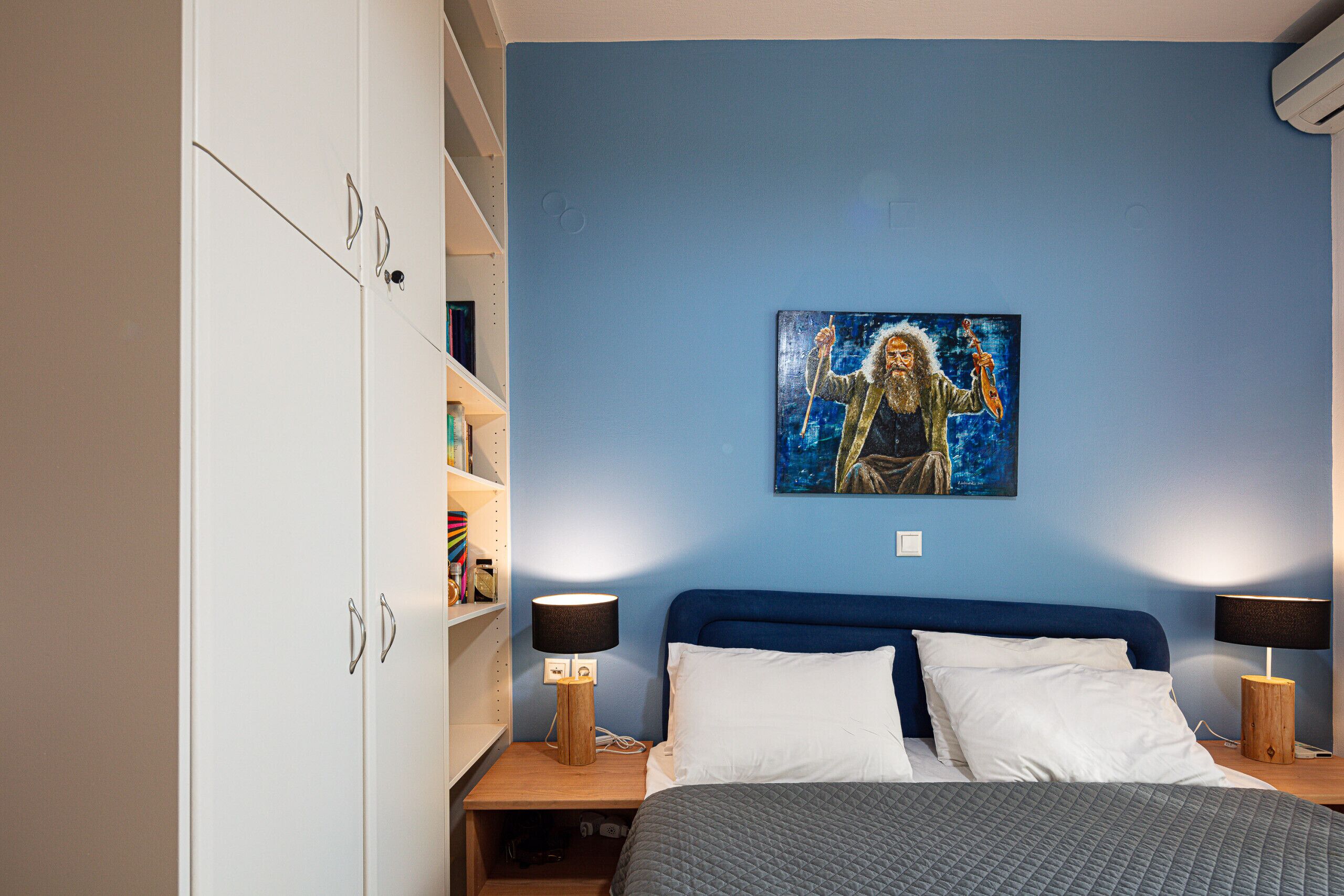 A bedroom with a blue wall, a bed with white and gray bedding, two bedside tables with lamps, and a painting above the bed.