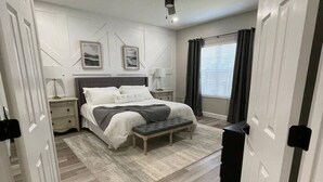 Ein Schlafzimmer mit einem Bett, Nachttischen, einer Bank, einem Deckenventilator und einem Fenster mit Vorhängen.