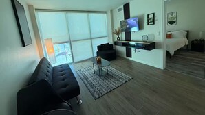 Ein modernes Wohnzimmer mit einer schwarzen Couch, einem Glastisch und einem flachbildfernseher, der an einer schwarzen Konsole befestigt ist. Ein großes Fenster bietet Blick ins Freie.