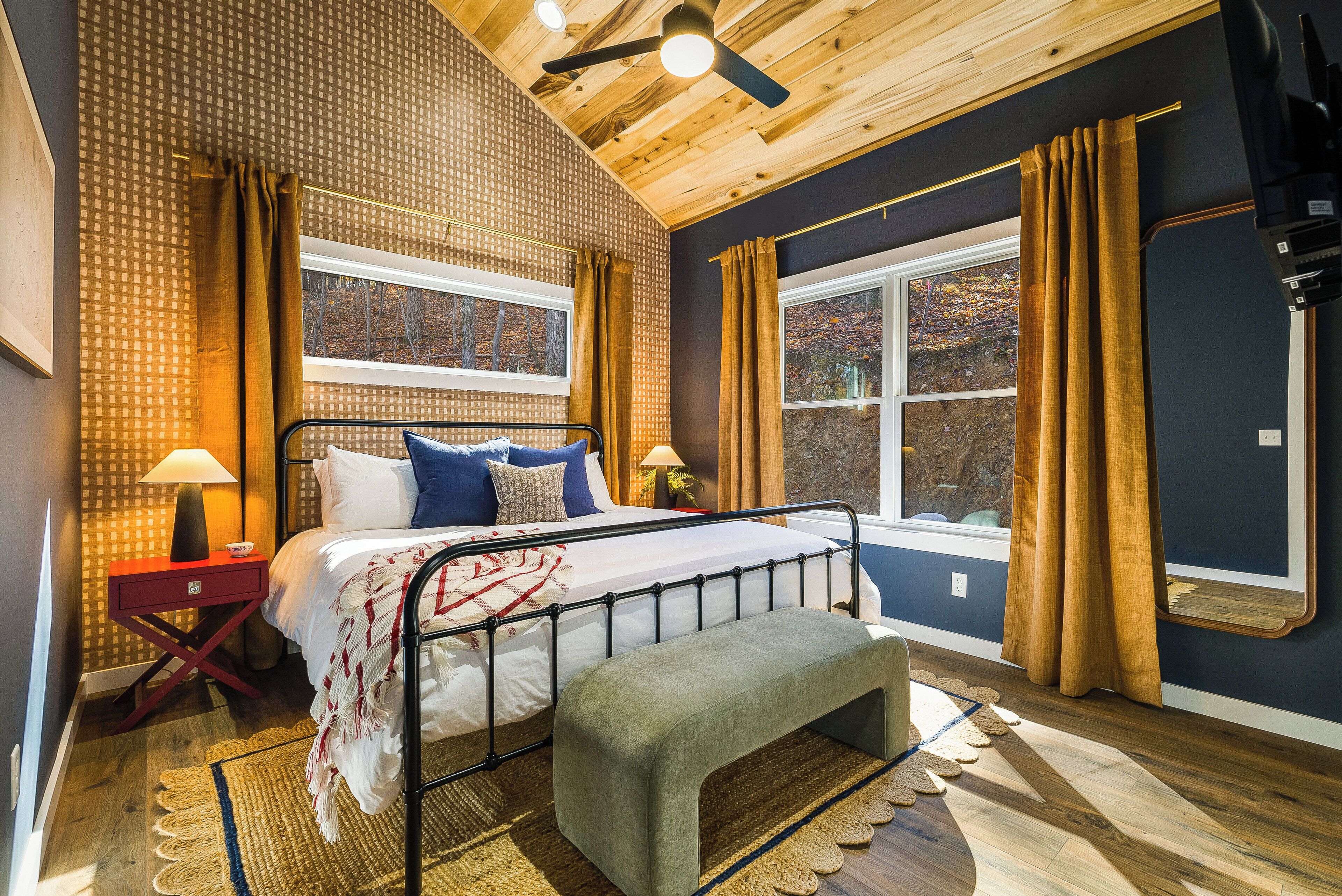 Un dormitorio con cama, mesitas de noche, un banco y ventanas amplias con cortinas.
