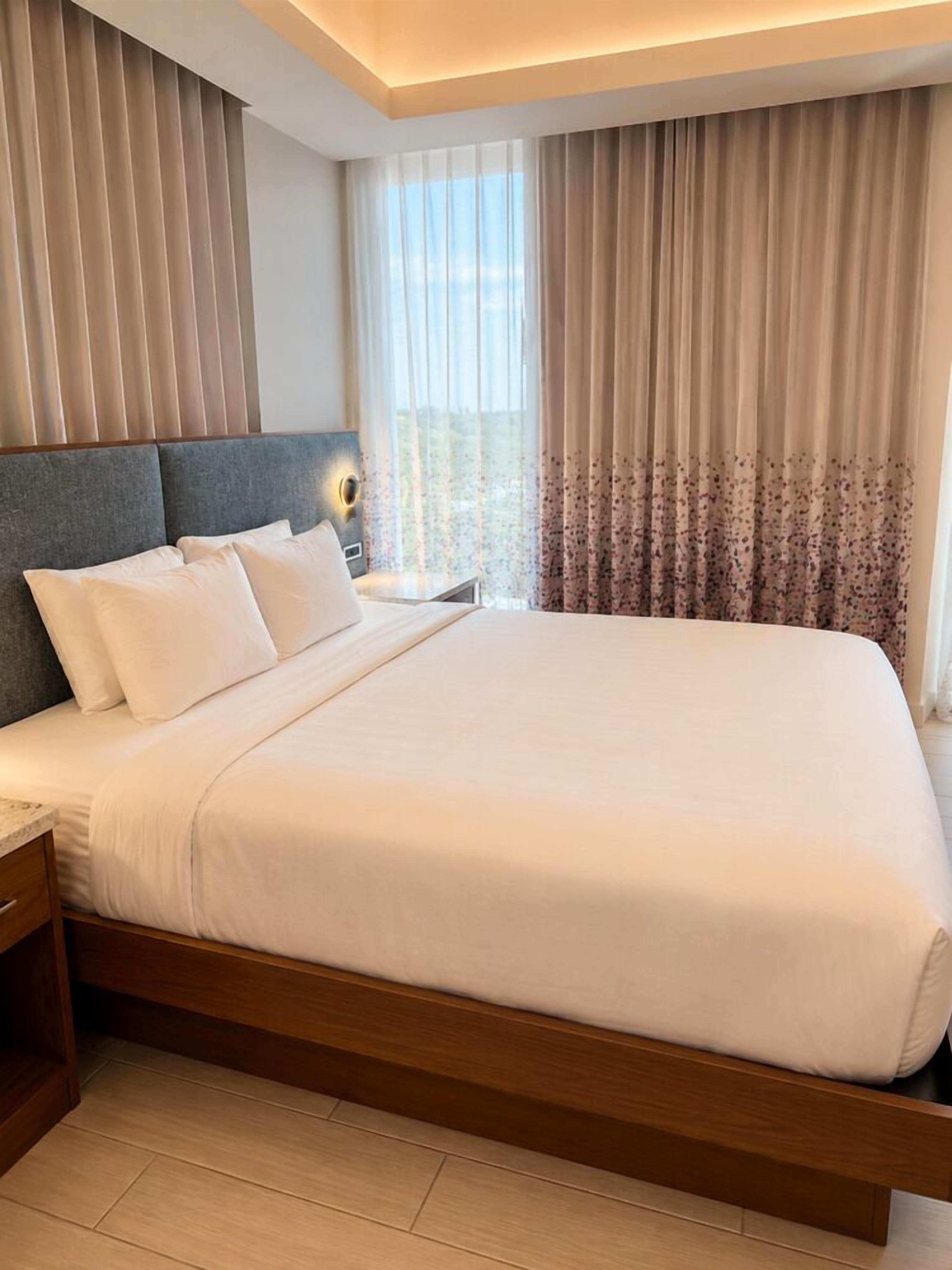Uma cama bem arrumada com cabeceira de madeira, mesa de cabeceira com abajur e janela com cortinas leves.