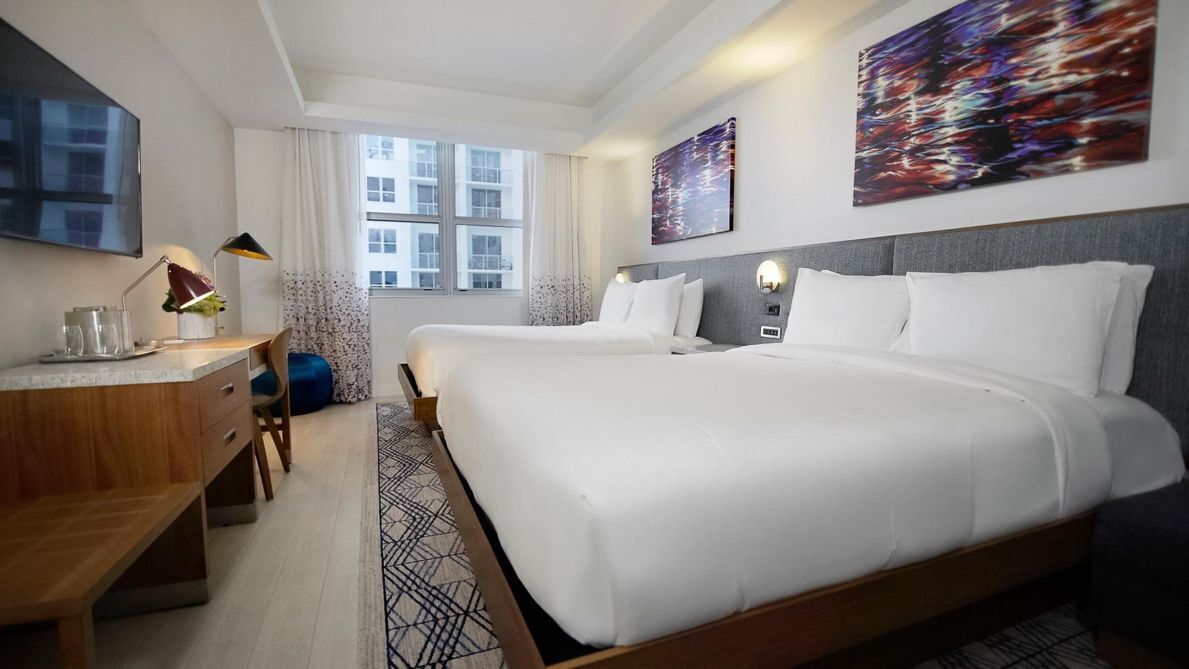 Ein Hotelzimmer mit zwei Betten, einem Schreibtisch, einem Stuhl, einem Fernseher und Bildern an den Wänden.