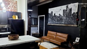 A modern kitchen with dark cabinets, a black refrigerator, and a microwave. There's a wooden bowl on the counter and a vase with sunflowers. A brown leather couch is in the living area with a cityscape painting on the wall. - Eclectic Loft in Historic Corktown where vibrance meets serenity – 75" TV & KING (Detroit)