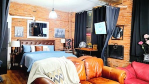 A room with a brick wall, a bed with a headboard, a leather sofa, a table with a lamp, and a window with curtains. - Eclectic Loft in Historic Corktown where vibrance meets serenity – 75" TV & KING (Detroit)