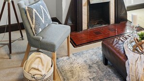 Une pièce avec une cheminée, un fauteuil, une table et une vue sur un bâtiment par la fenêtre.
