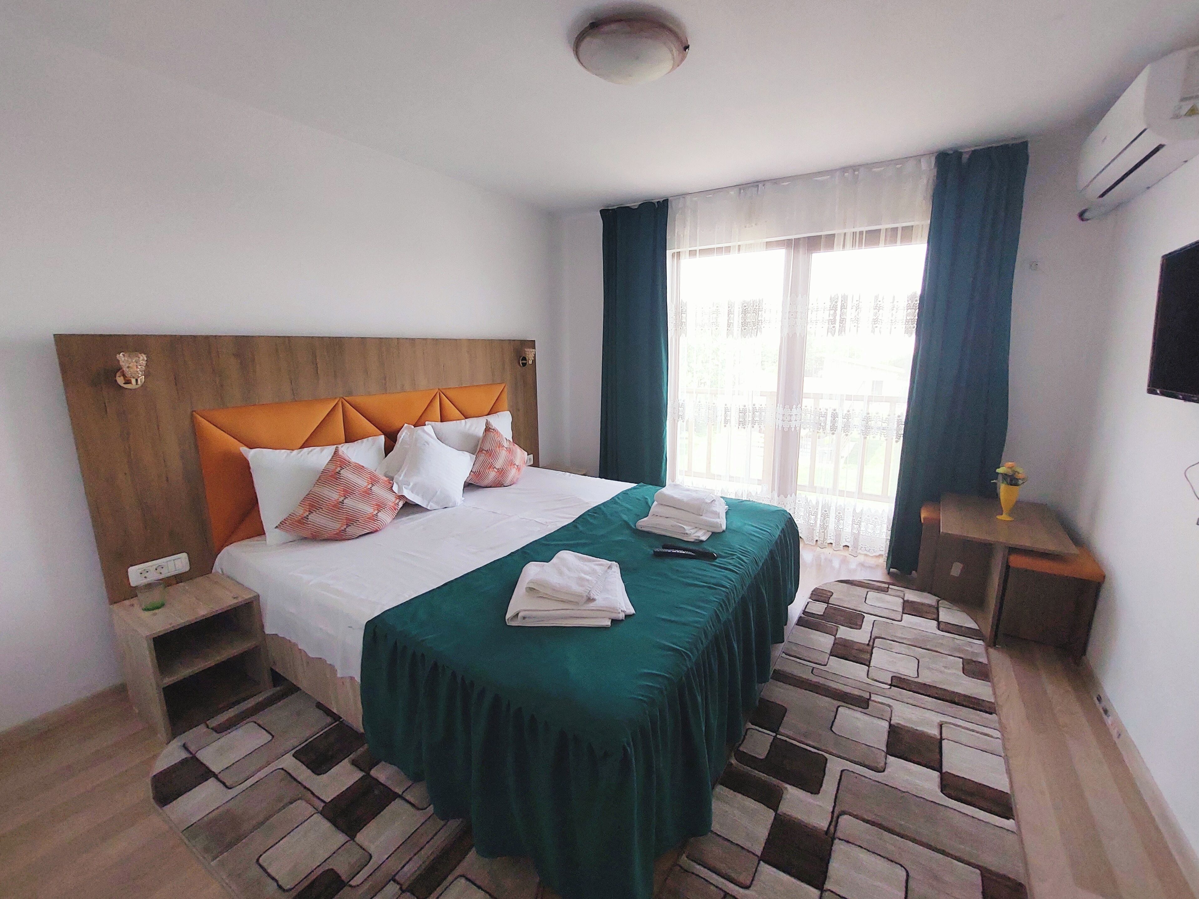 Ein Hotelzimmer mit einem hölzernen Kopfteil, einem Bett mit grün-weißer Bettwäsche, einem kleinen Nachttisch und einem an der Wand befestigten Fernseher.