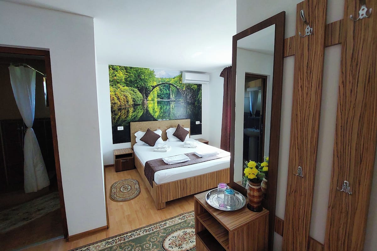 A bedroom with a bed, a mirror, a bedside table with a vase of flowers, and a view of a garden through the window.