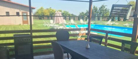 Un patio techado con mesa y sillas, piscina al fondo y vista a un área con césped.