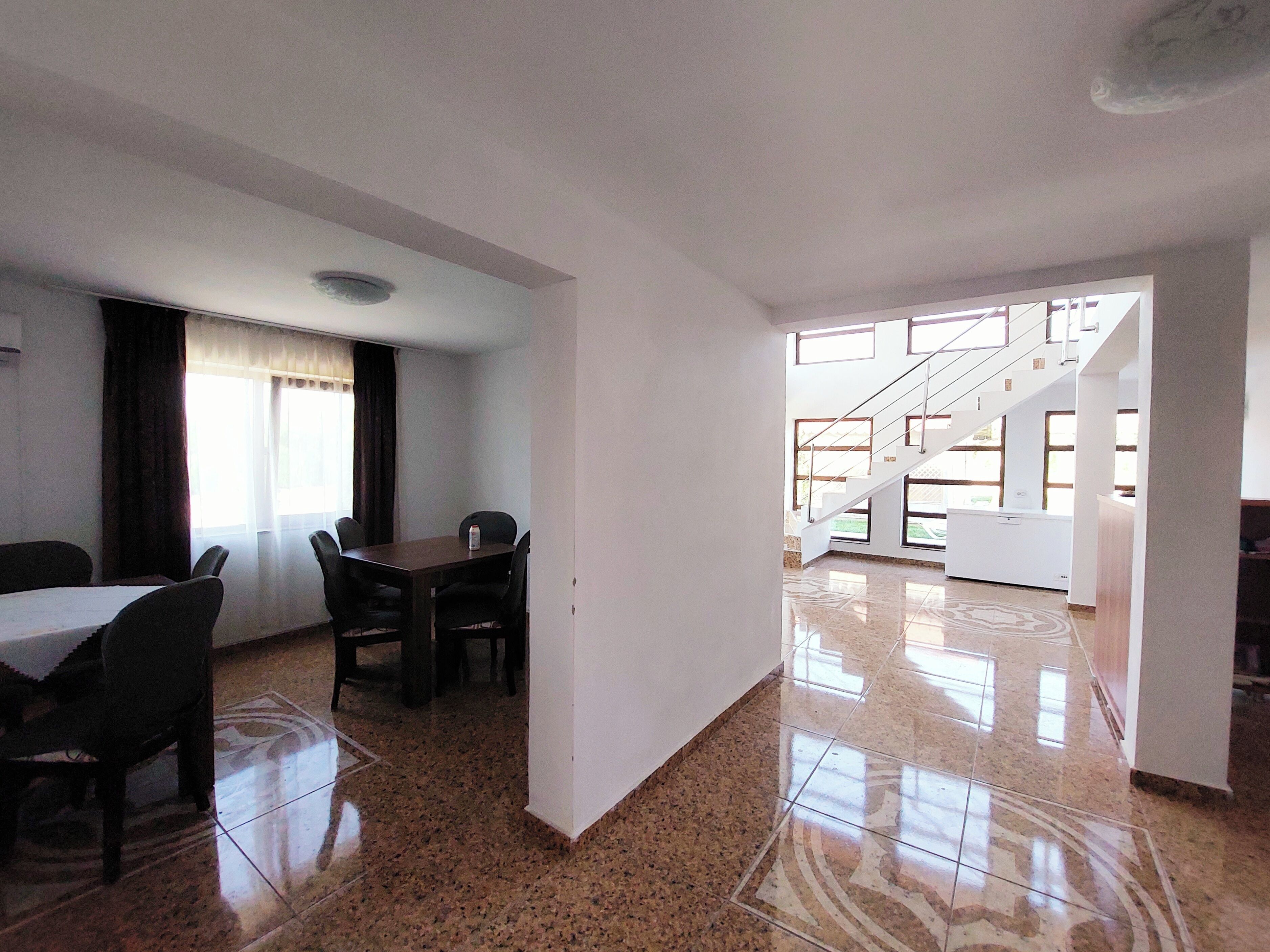 A spacious room with a dining area, a staircase, and large windows.