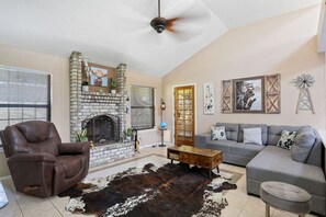 A living room with a brick fireplace, a leather armchair, a grey sofa, and a cowhide rug. - Casa Mi Ranchito Alegre (El Paso)