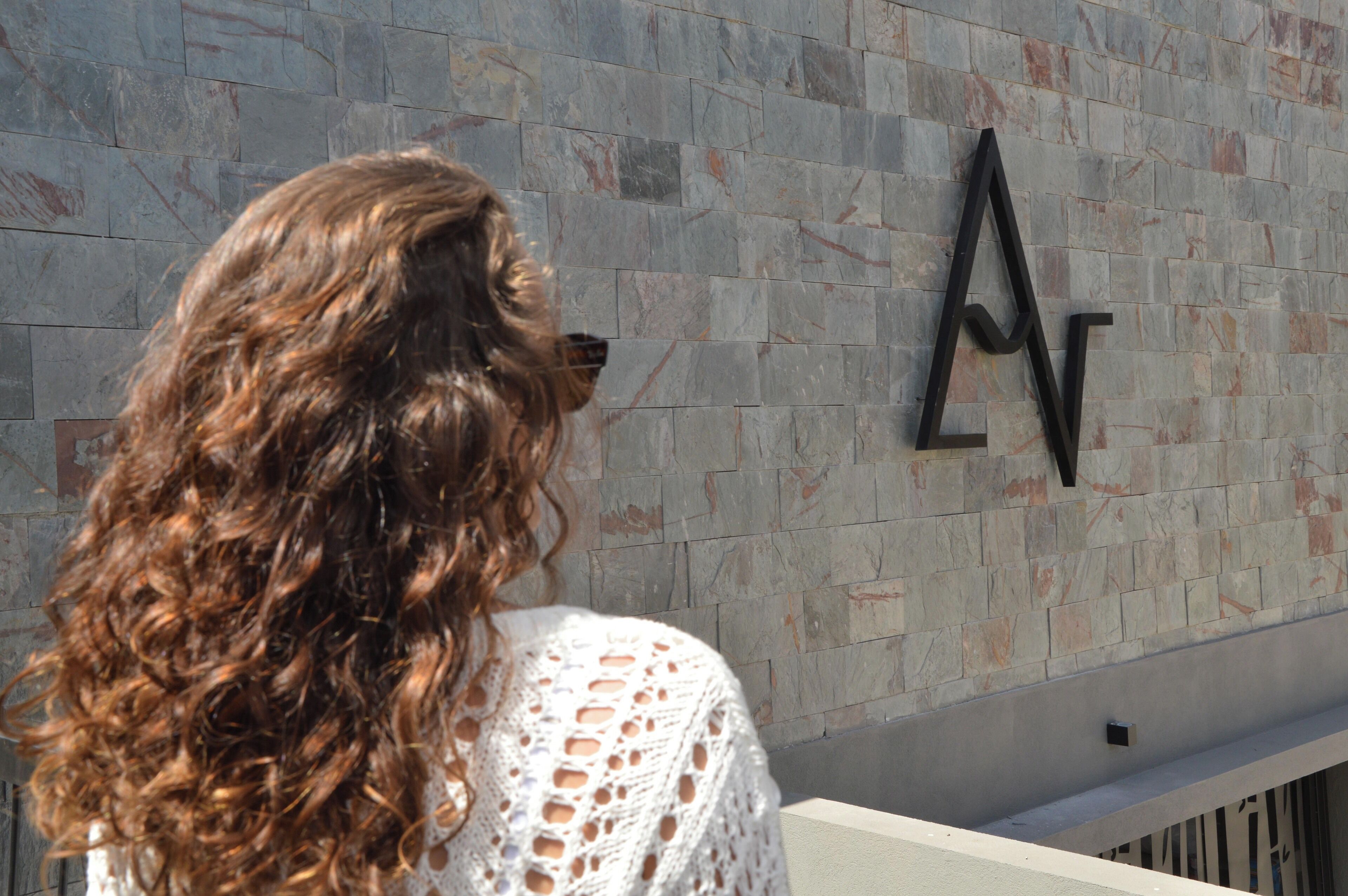 A person with long curly hair standing in front of a wall with a geometric design.