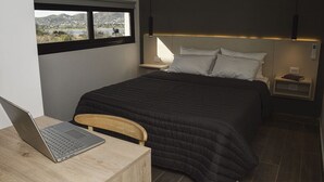 Een slaapkamer met een bed, een bureau met een laptop, een stoel, een nachtkastje met een lamp, en uitzicht op een landschap.