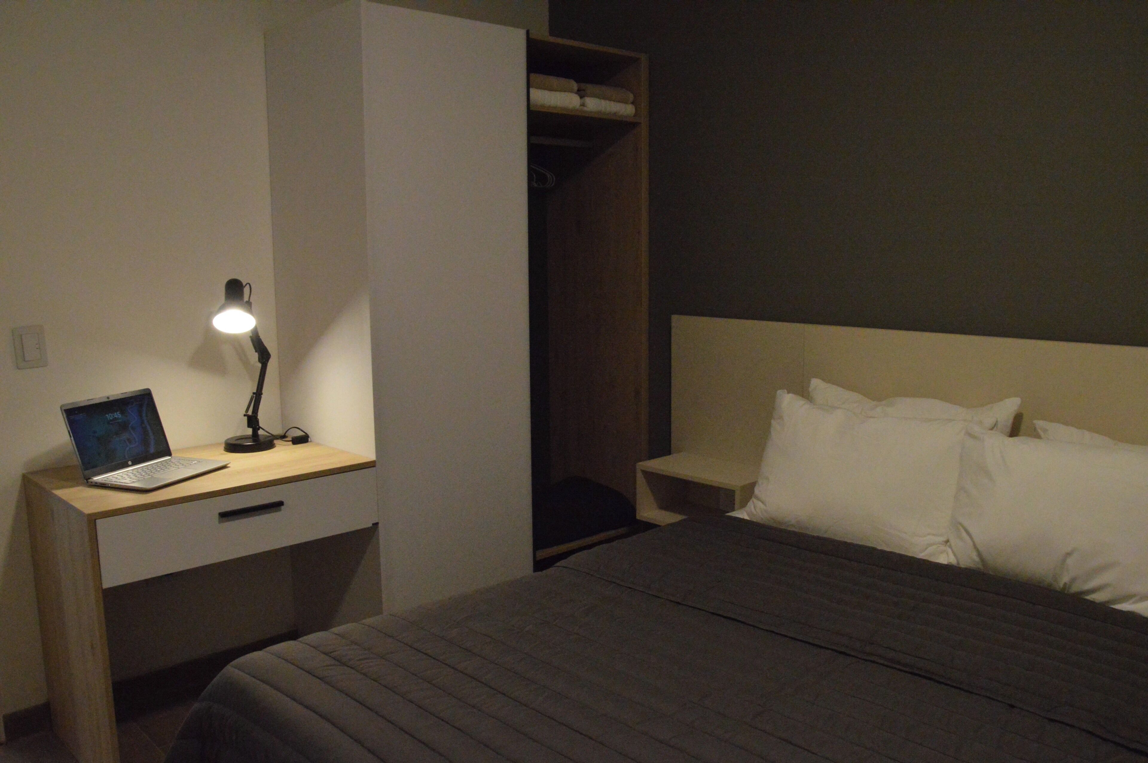 A hotel room with a bed, a desk with a laptop, a bedside table with a lamp, and a wardrobe.
