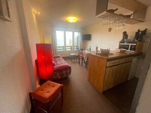 Un espacio habitable compacto con kitchenette, comedor y zona de estar con sofá y reposapiés.