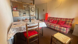 A cozy living room with a dining area. There is a sofa with red cushions, a dining table set with plates and glasses, and a clock on the wall. - Pra-Loup 1600 - T2 Apartment for 6, central location, near slopes (Pra-Loup)