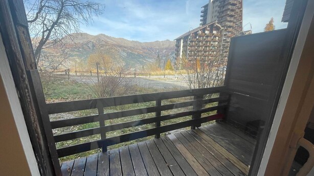 A balcony with a wooden floor and metal railing, overlooking a mountainous landscape and a multi-story building. - Pra-Loup 1600 - T2 Apartment for 6, central location, near slopes (Pra-Loup)