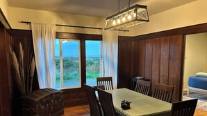 A dining area with a table and chairs, a hanging light fixture, and a view through large windows. - Oceanview Retreat — 2BR, Walk to the Beach (Samoa)