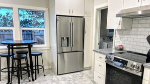 A modern kitchen with a stainless steel refrigerator, a gas stove, and white cabinetry. There is a round table with stools and a window with a view of trees. - Oceanview Retreat — 2BR, Walk to the Beach (Samoa)