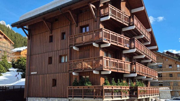 Ein mehrstöckiges Holzgebäude mit Balkonen, einem Steinfundament und einer Skipiste im Hintergrund.