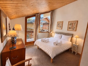 Ein Schlafzimmer mit Holzboden, einem Bett mit weißen Leinen, zwei Nachttischlampen, einer Holzkommode und einem Fenster mit Blick auf ein Holzhaus.