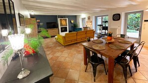 Een ruime woonkamer met een bank, eettafel en stoelen. Achterin is een televisie en een open haard te zien.