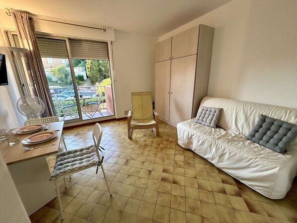 A small, well-lit room with a dining table, chairs, a sofa, and a large window with blinds. - Cozy Studio with Balcony near the Port - Cap d'Agde (Cap d'Agde)