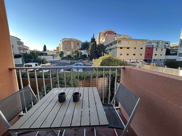 A balcony with a wooden table and two chairs, overlooking a parking area and buildings.