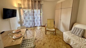 A room with a dining table set for two, a sofa, a chair, a TV mounted on the wall, and a wardrobe.
