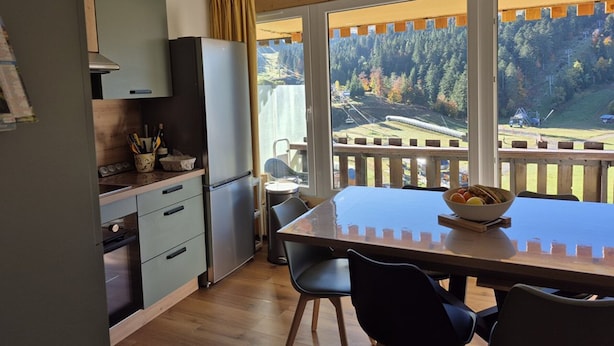 Cozinha moderna com área de refeições, janelas amplas com vista para uma floresta e uma ponte, e teto de madeira.