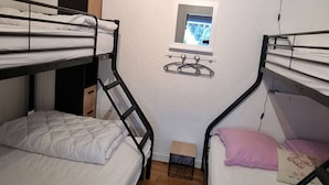 Quarto em dormitório com beliches, uma janela pequena, uma prateleira com um ursinho de pelúcia e um banquinho de madeira.