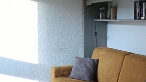 Ein Wohnzimmer mit einem senfgelben Sofa, einem grauen Kissen und einem Bücherregal mit Büchern und Dekorationselementen.