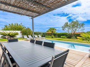 A poolside area with a table and chairs, a covered patio, and a view of the pool and greenery.