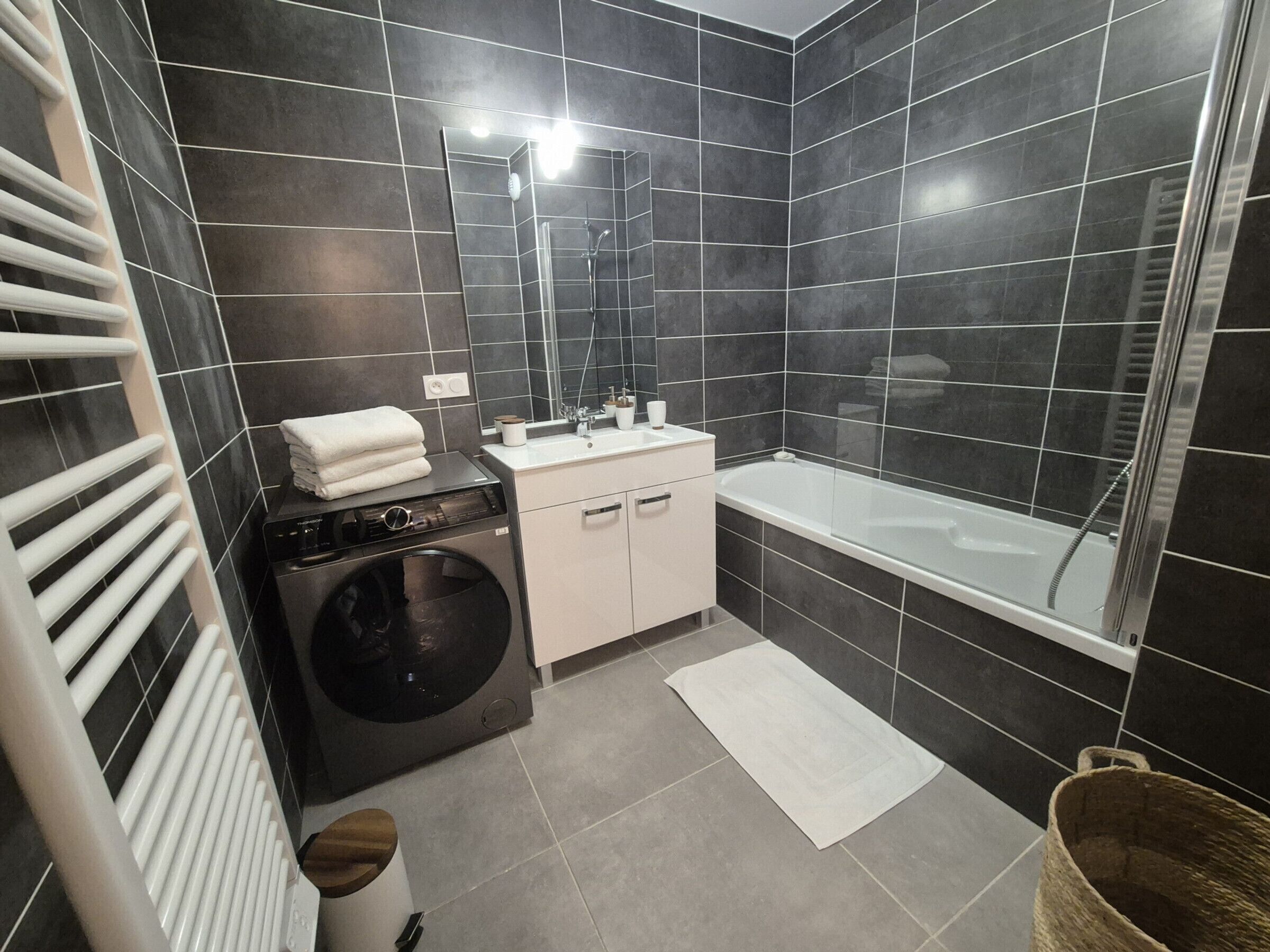Une salle de bain moderne avec un mur recouvert de carreaux noirs, un lavabo blanc, une baignoire, un lave-linge et un porte-serviettes blanc.