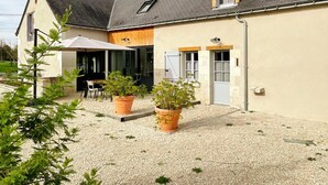 Ein Haus mit Kiesweg, einer überdachten Terrasse und Gartenmöbeln, sowie Kübelpflanzen.