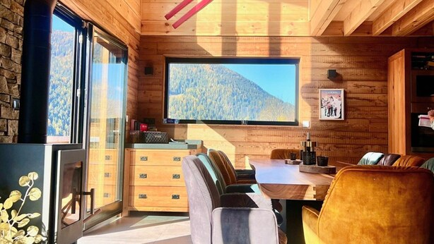 Interior acogedor de una cabaña con detalles de madera, un ventanal grande que ofrece vistas a la montaña, un comedor con mesa y sillas, y una chimenea.