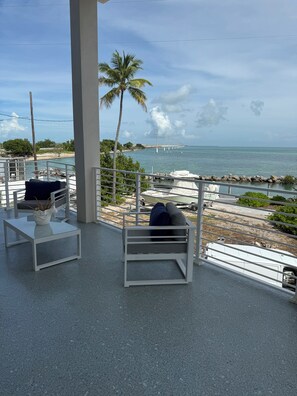 Ein Balkon mit weißer Metallreling, einem weißen Tisch mit Topfpflanze und einer gepolsterten Bank. Im Hintergrund sind Strand, Steg und Ozean zu sehen.