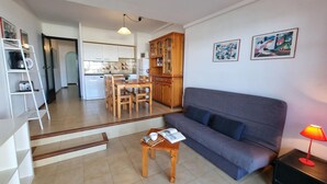 A living room with a grey sofa, a wooden coffee table with a magazine, and a red lamp. There is a staircase leading to a kitchen with white cabinets and a dining area with wooden chairs. - Apartment in L'Escala with terrace near the beach (L'Escala)