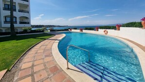 En swimmingpool med ett kaklat poolområde, omgiven av en vit byggnad med balkonger, och en fri utsikt över havet i bakgrunden.