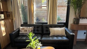 A living room with a black leather sofa, a wooden coffee table, and a large window with curtains. - The Roncy Den ~a boutique laneway house (Toronto)