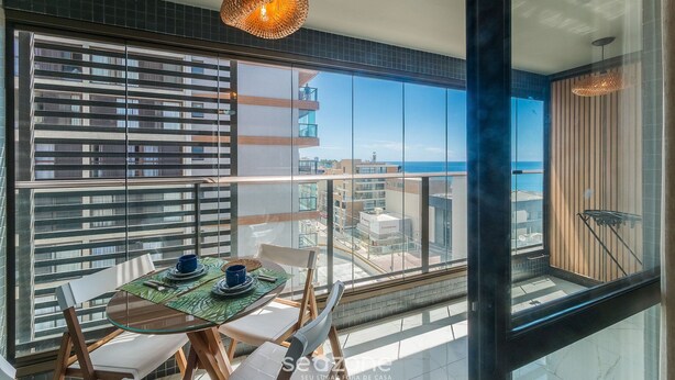A modern balcony with a glass table, chairs, and a hanging light fixture. - Apts 120 m From Barra Beach With Pool and gym in the Building - Esbs (Salvador)