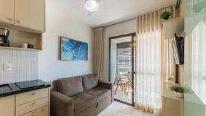 A modern kitchen with a black countertop, beige cabinets, and a grey sofa. There is a painting on the wall and a window with curtains. - Apts 120 m From Barra Beach With Pool and gym in the Building - Esbs (Salvador)