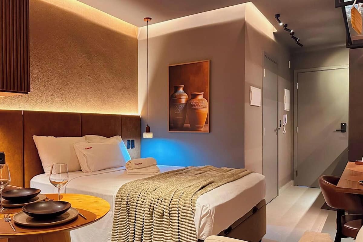 Quarto de hotel moderno com cama, mesa de cabeceira e um espaço de refeições com mesa e cadeiras.