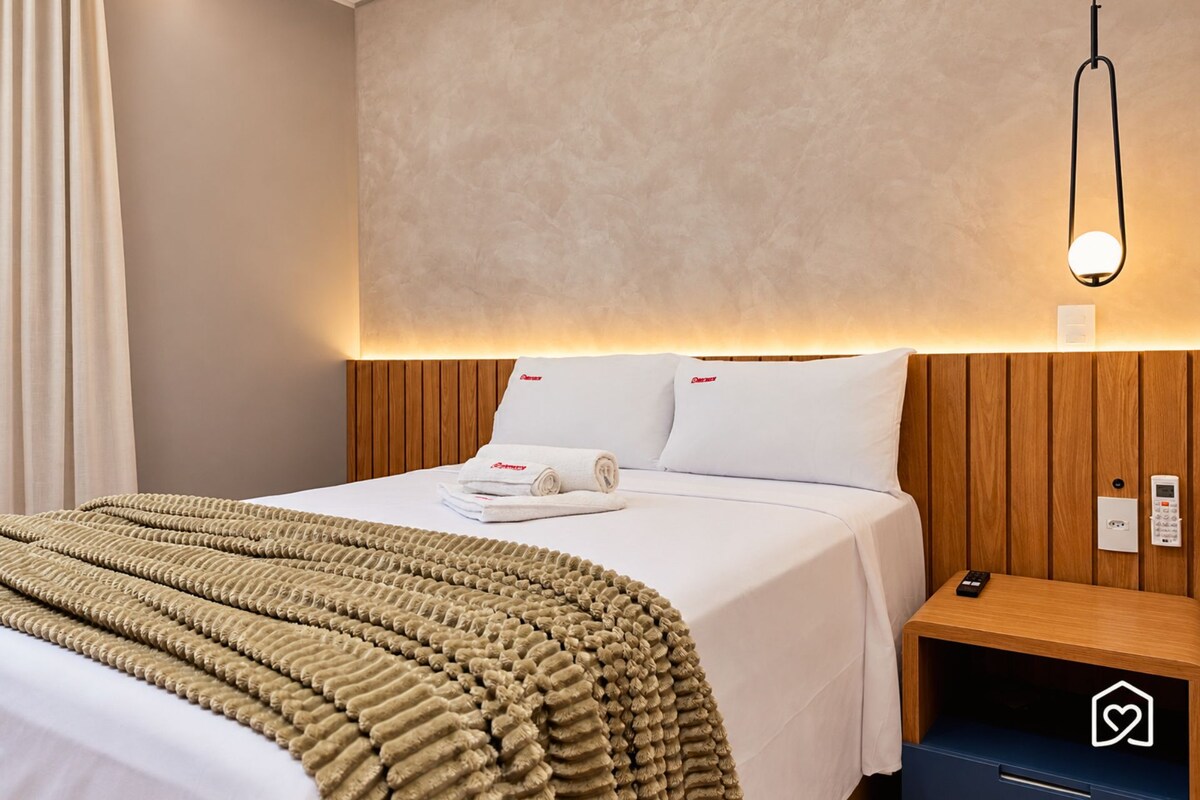 A hotel room with a bed, a bedside table with a remote control, and a wall-mounted light fixture.