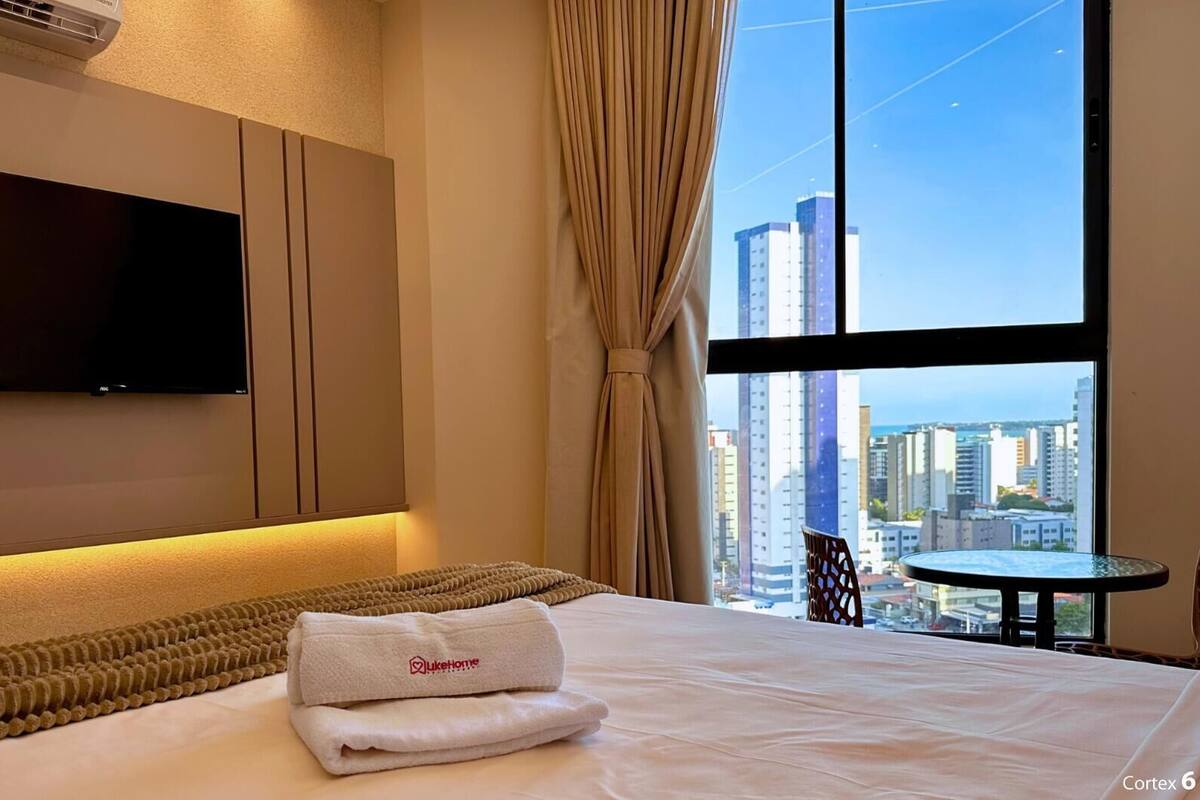 Quarto de hotel com cama, televisão de tela plana, mesinha e vista para a paisagem urbana.