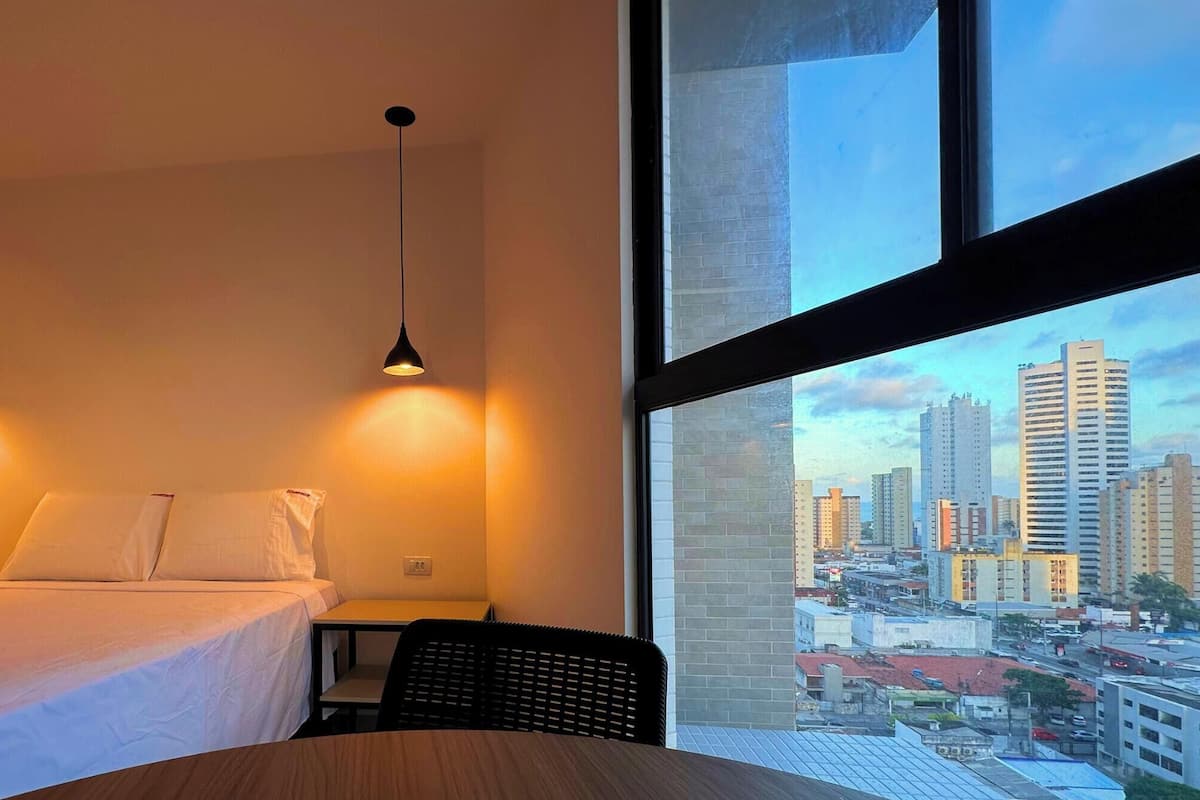 Quarto de hotel com cama, uma pequena mesa, duas luzes pendentes e uma janela grande com vista para a cidade.