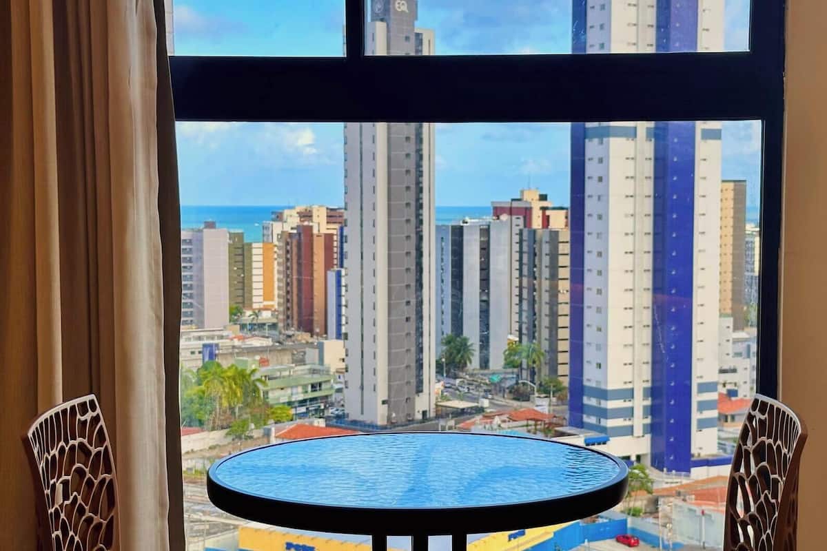 Vista da janela com uma mesa redonda e cadeiras, mostrando um horizonte urbano com edifícios altos e céu claro.