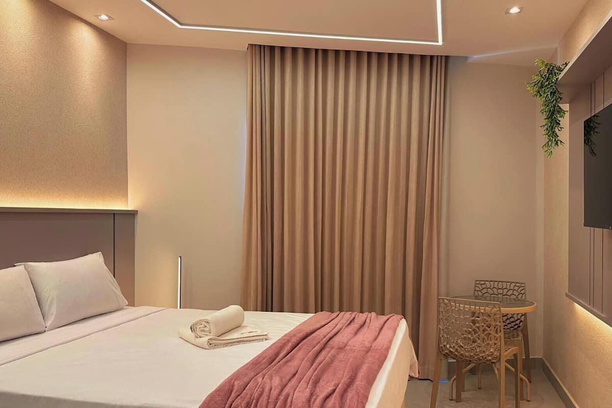 Quarto de hotel moderno com cama bem arrumada, uma pequena mesa com cadeira, TV fixada na parede e uma janela grande com cortinas.