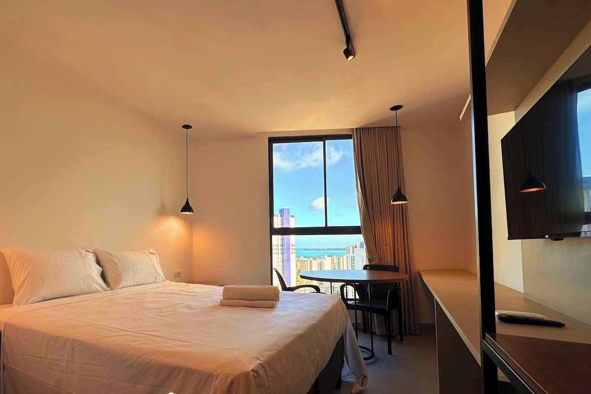 Quarto de hotel com cama, mesas de cabeceira, escrivaninha, cadeira e televisão.