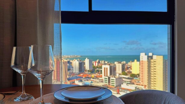 Mesa com duas taças de vinho e um prato, à frente de uma janela com vista para uma paisagem urbana com edifícios e um corpo de água.